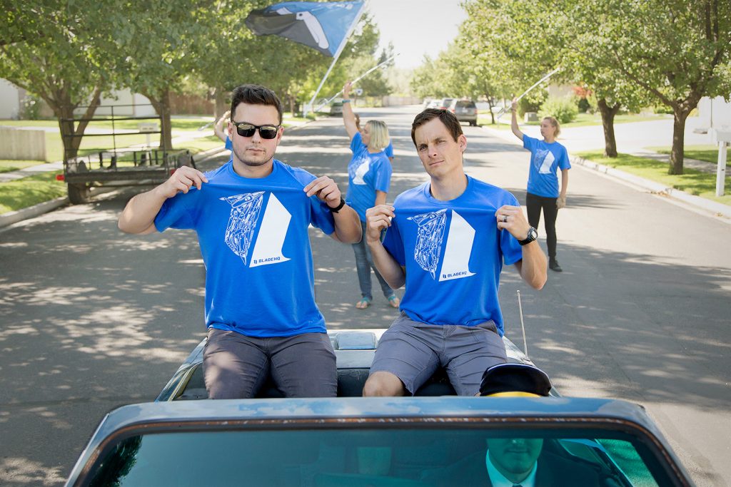 Three men in a car indicating their Blade HQ shirts, with three women behind the car waving a flag, all celebrating National Knife Day.