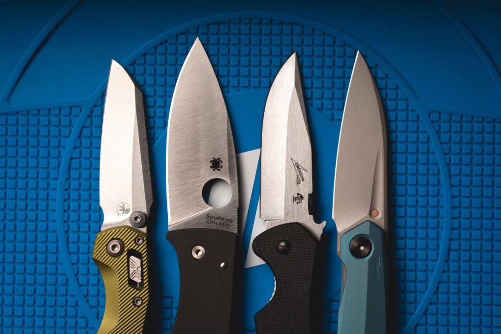 Four knives side by side on a blue Blade HQ shop mat. Each has a different opener: thumb stud, thumb hole, thumb disk, and thumb groove.