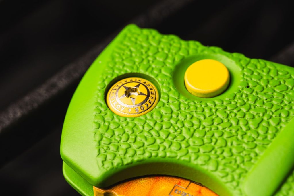 Closeup of the Boker KalashniCob showcasing the pivot screw with Boker's branding, the yellow automatic switchblade button lock, and the husky texture of the green handle.