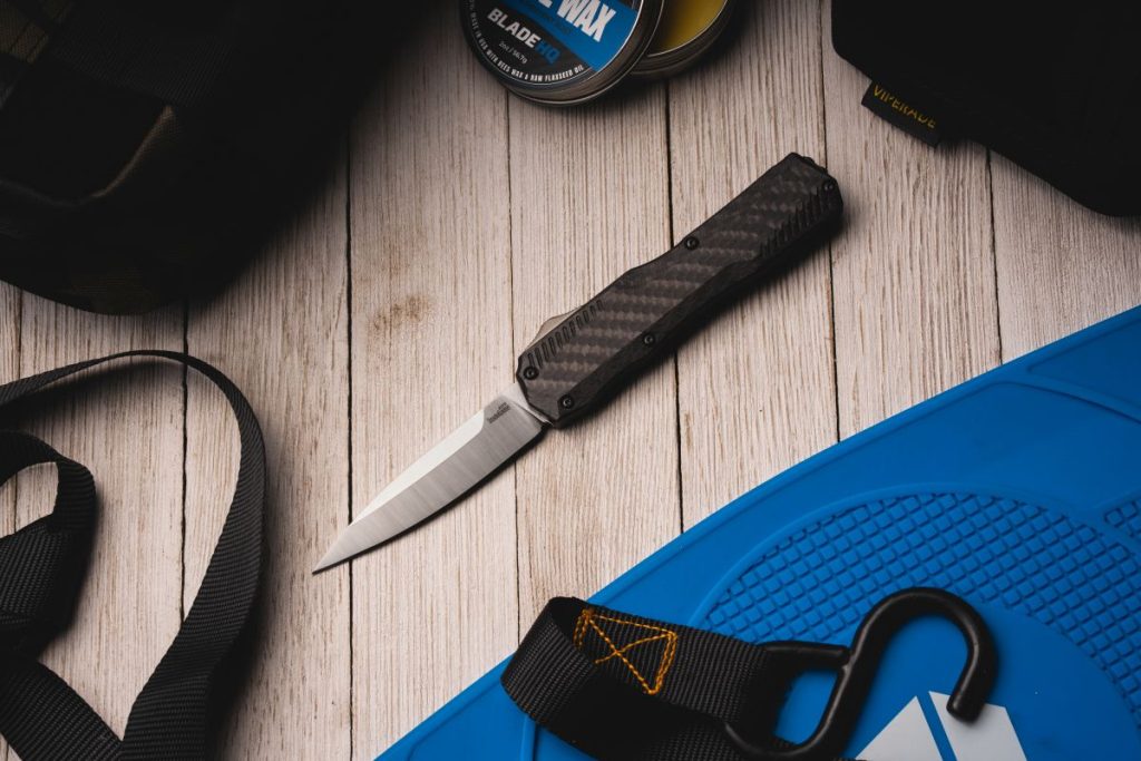 A Kershaw Livewire OTF knife with a carbon fiber handle on a table next to gear.