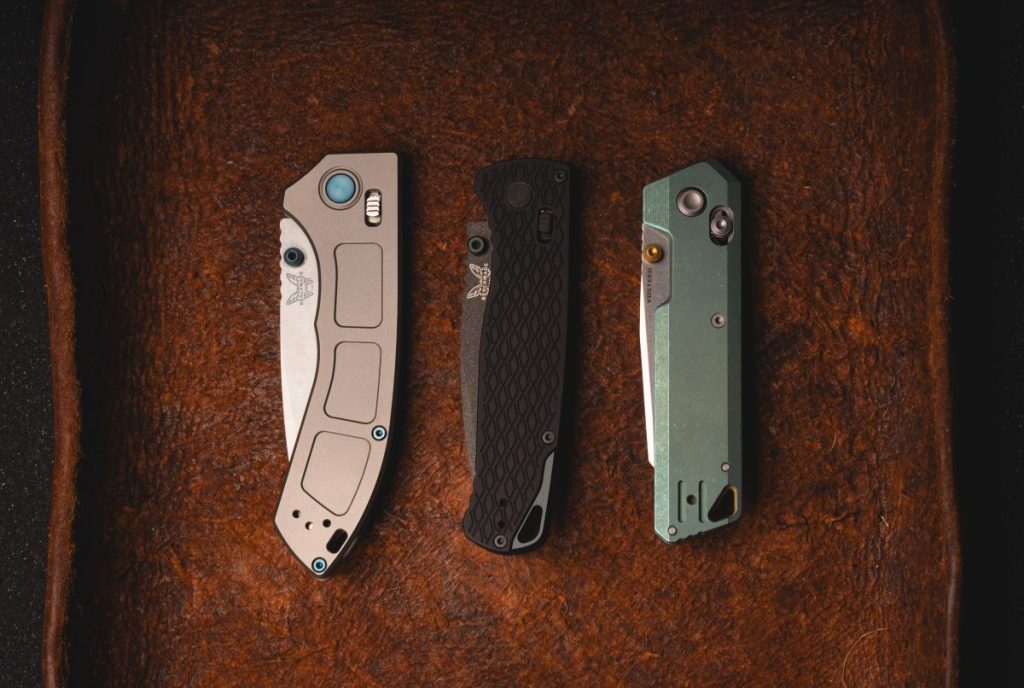 The Benchmade Narrows (gray), Benchmade Bugout Vapyr (black), and Vosteed Parallel (green) in a row on a leather mat.