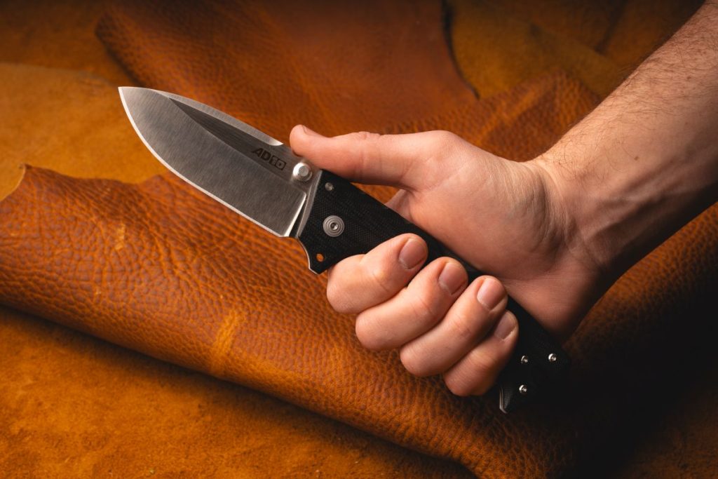 A man holds a Cold Steel AD-10, which features the ultra-strong Tri-Ad lock. The background is leather.
