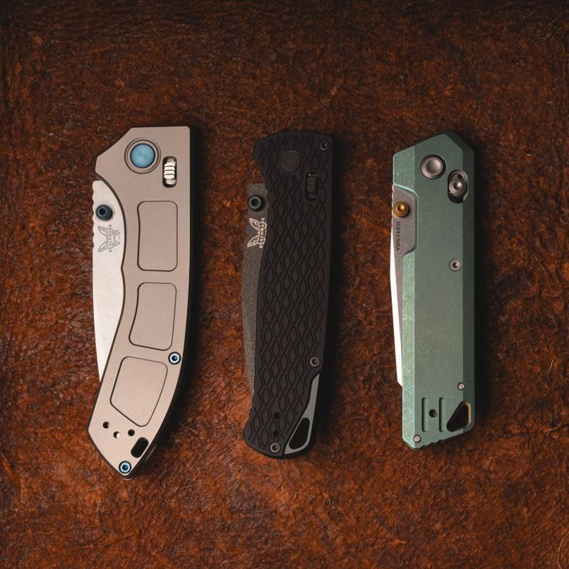 The Benchmade Narrows (gray), Benchmade Bugout Vapyr (black), and Vosteed Parallel (green) in a row on a leather mat.